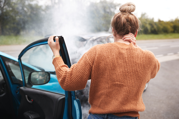 A young woman with her back to the camera stands outside a passenger side door that is open and holds her other hand to her hurting neck while looking at the car they crashed into in front of her