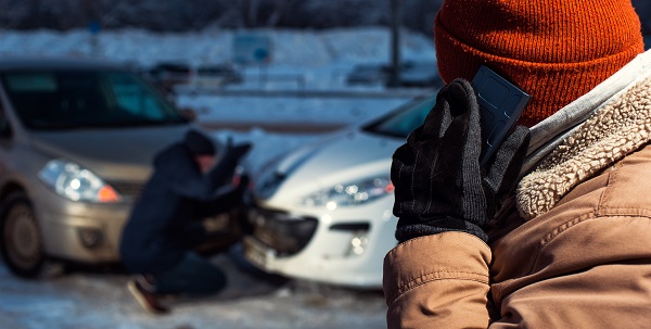 Man in the foreground in winter clothes making a phone call because he has crashed car with another driving who is kneeling next to the cars in the background