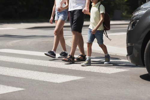 A father holding the hands of his two children crosses in a crosswalk in front of a stopped car