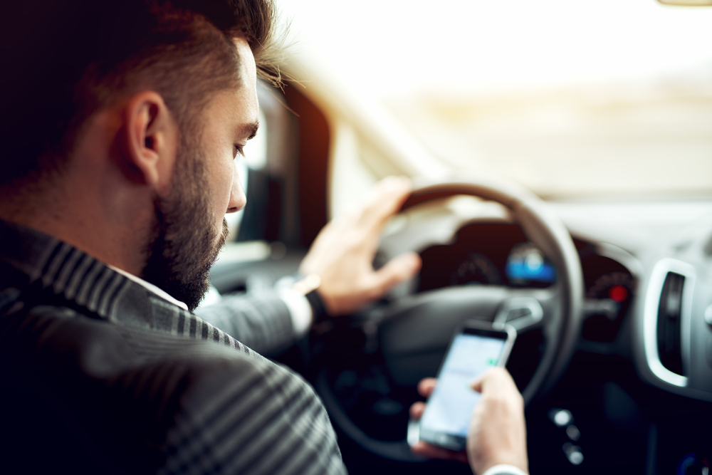 A man is looking at his cell phone screen with one hand on the steering wheel
